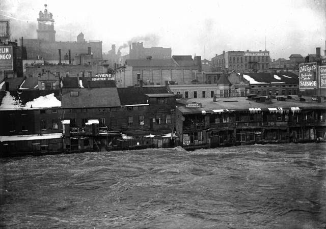 Genesee River in high water stage, showing the backs of Front Street buildings. This is believed to be a depiction of the flood of 1913. Myer's Department Store, Charles Adam (grinding), and Zweigle's (sausage maker and saloon) are visible. Weis & Fisher Company (furniture and carpets) is in the right background at 297 Brown Street. The Powers Building tower is in the upper left corner, 1913