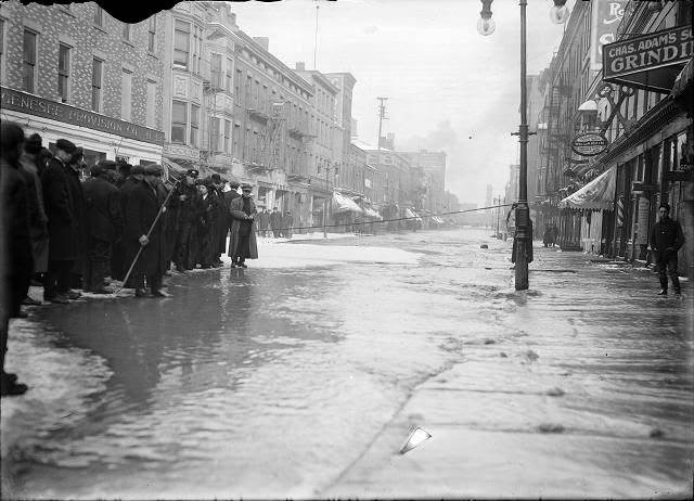 People stand at the edge of the flood water on Front Street in front of the Genesee Provision Company. A rope cordons off the street near Charles Adam's Grinding and the William H. Larkin Hotel, 1913