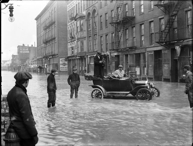 Two men are stopped in a Ford car during the flood on Front Street. One man is holding a camera and other men stand in the street in ankle-deep water, 1913