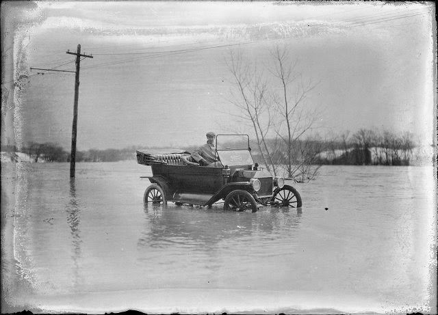 A man sits in a Ford automobile, which has water halfway up the wheels. This is Plymouth Avenue, which has been completely flooded by the Genesee River, 1913