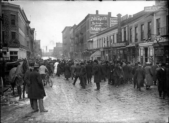 People, including one man with a horse and buggy, stand at the edge of the flood on Front Street. Dave Solomon, Pawnbroker and Auctioneer; Charles Adam's Sons, Incorporated, Grinding; Zweigle's; and (probably) the Genesee Provision Company are visible, 1913