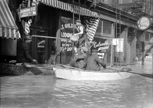 Men climb up a ladder from a rowboat to deliver food to families marooned on the upper floors of Rochester's business section. The men are pictured in front of Jacob Goldstein's Lodging House and J.C. Vine's Columbia Hotel, 1913