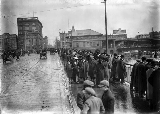 Pedestrians throng the sidewalks of the Court Street Bridge to see the flood waters, as a few horse-driven carriages and automobiles drive past, 1913