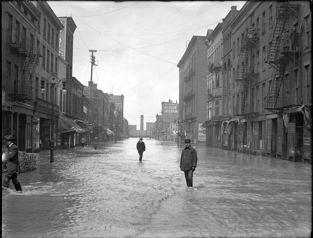 Men are standing ankle-deep in water on Front Street. On the left is the Atlantic Supply House and on the right is the People's Rescue Mission, 1913
