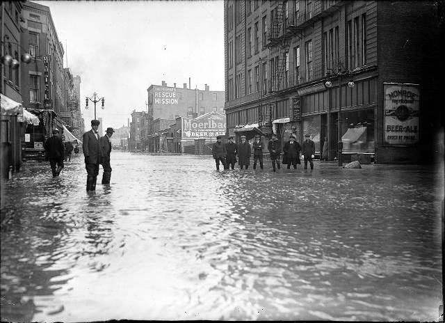 Men stand ankle deep in flood water on Front Street. They are in front of the Rochester Poultry & Commission Company. The People's Rescue Mission is in the background, 1913