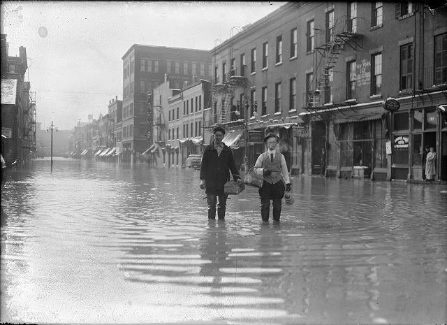 Two men stand ankle-deep in water on Front Street. They are carrying a coffee pot and baskets. On the right a man and a woman look out of a doorway, 1913