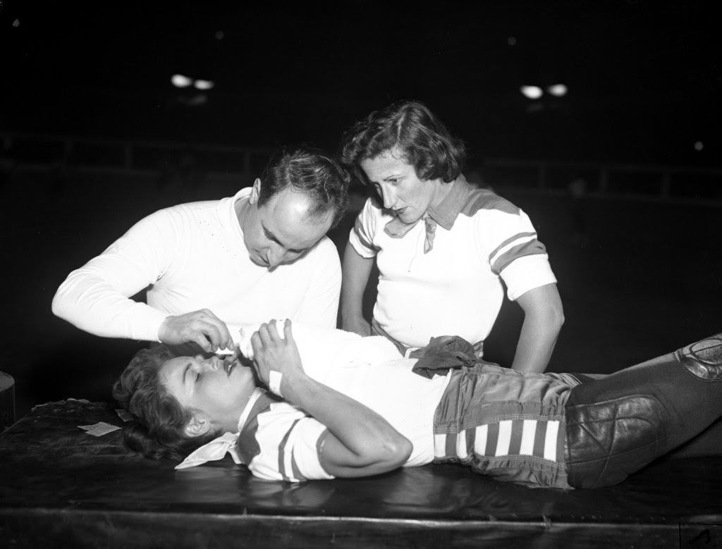 Betty Boyd of the Jersey team is treated by Dr. James Alvian as her teammate and captain Annis Jansen watches at a roller derby at the Coliseum, 1950s